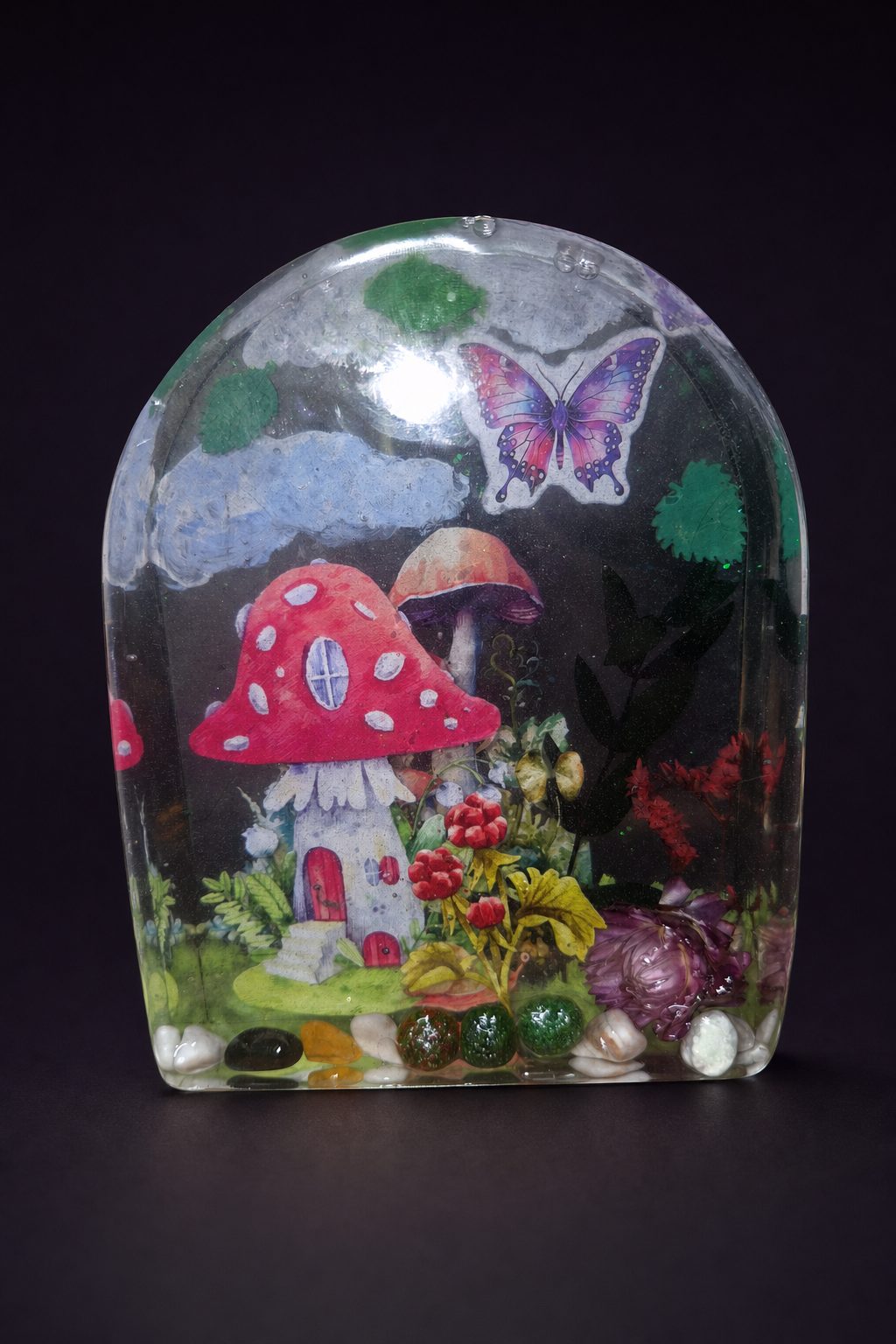 Arched resin still life sculpture with a mushroom house, clouds, butterfly, real flowers, and crystal chips on a dark plum background