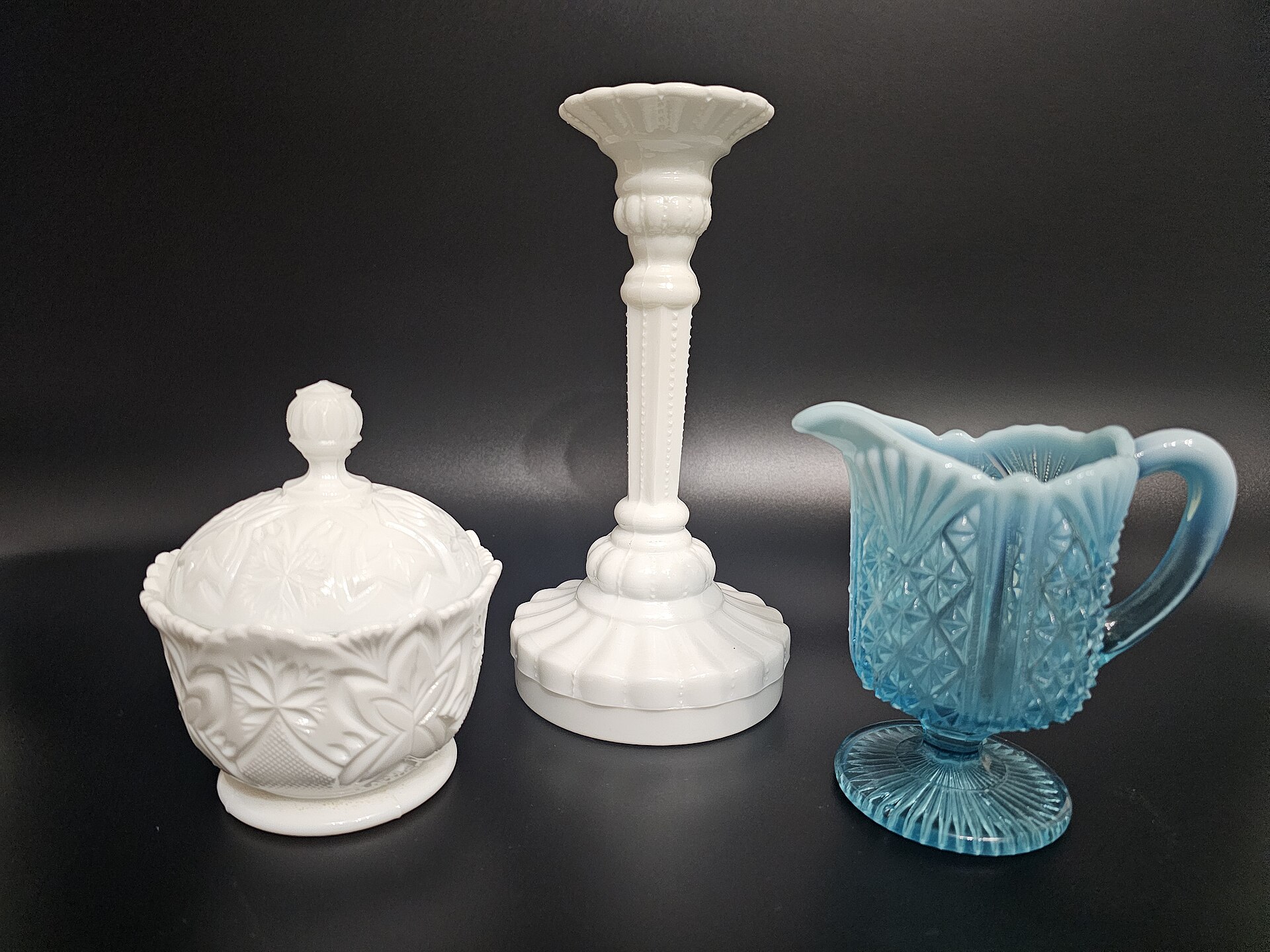 White milk glass sugar bowl and candlestick shown beside blue pressed glass