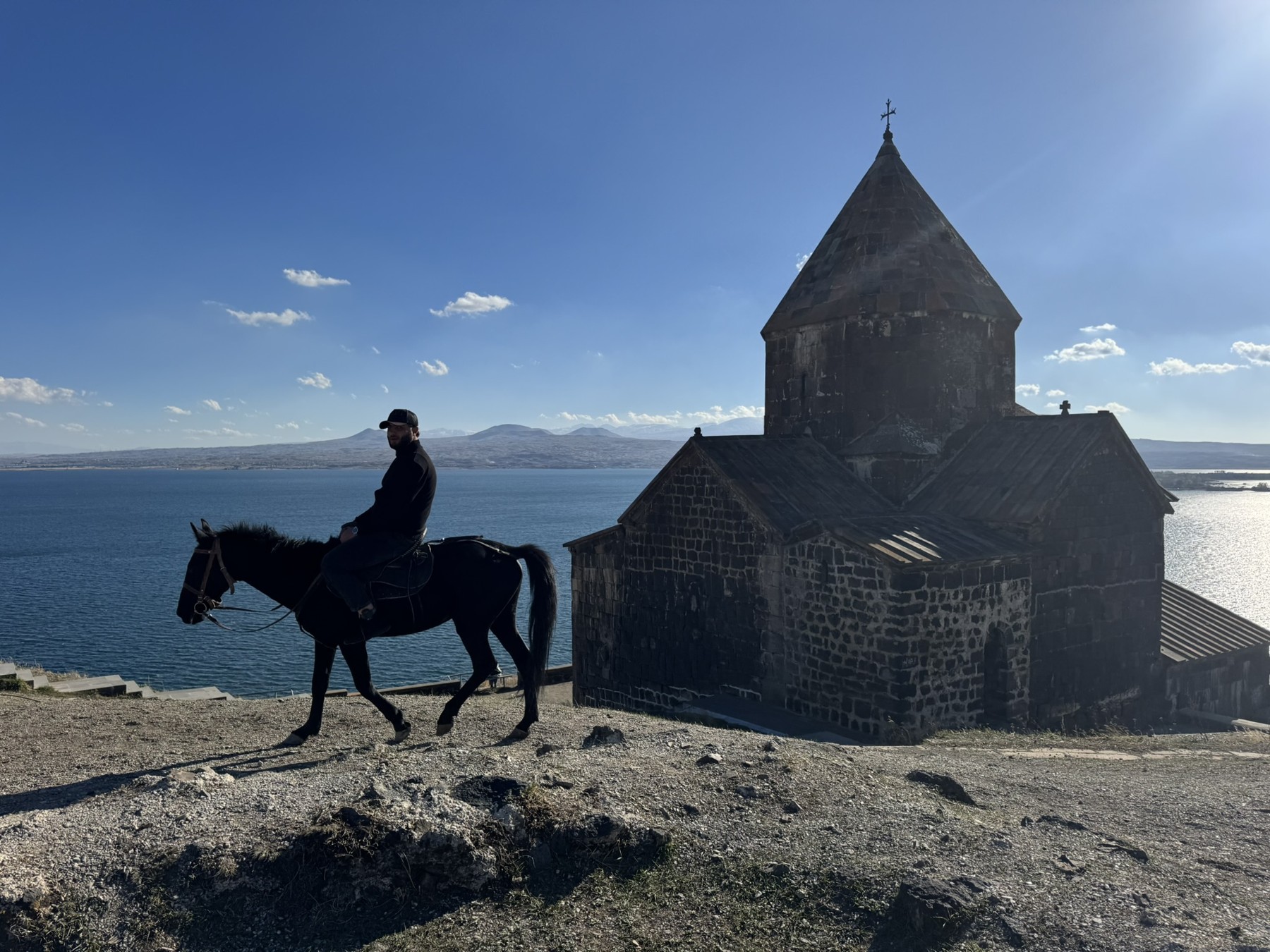 A rider on a dark horse beside a hilltop church above Lake Sevan.