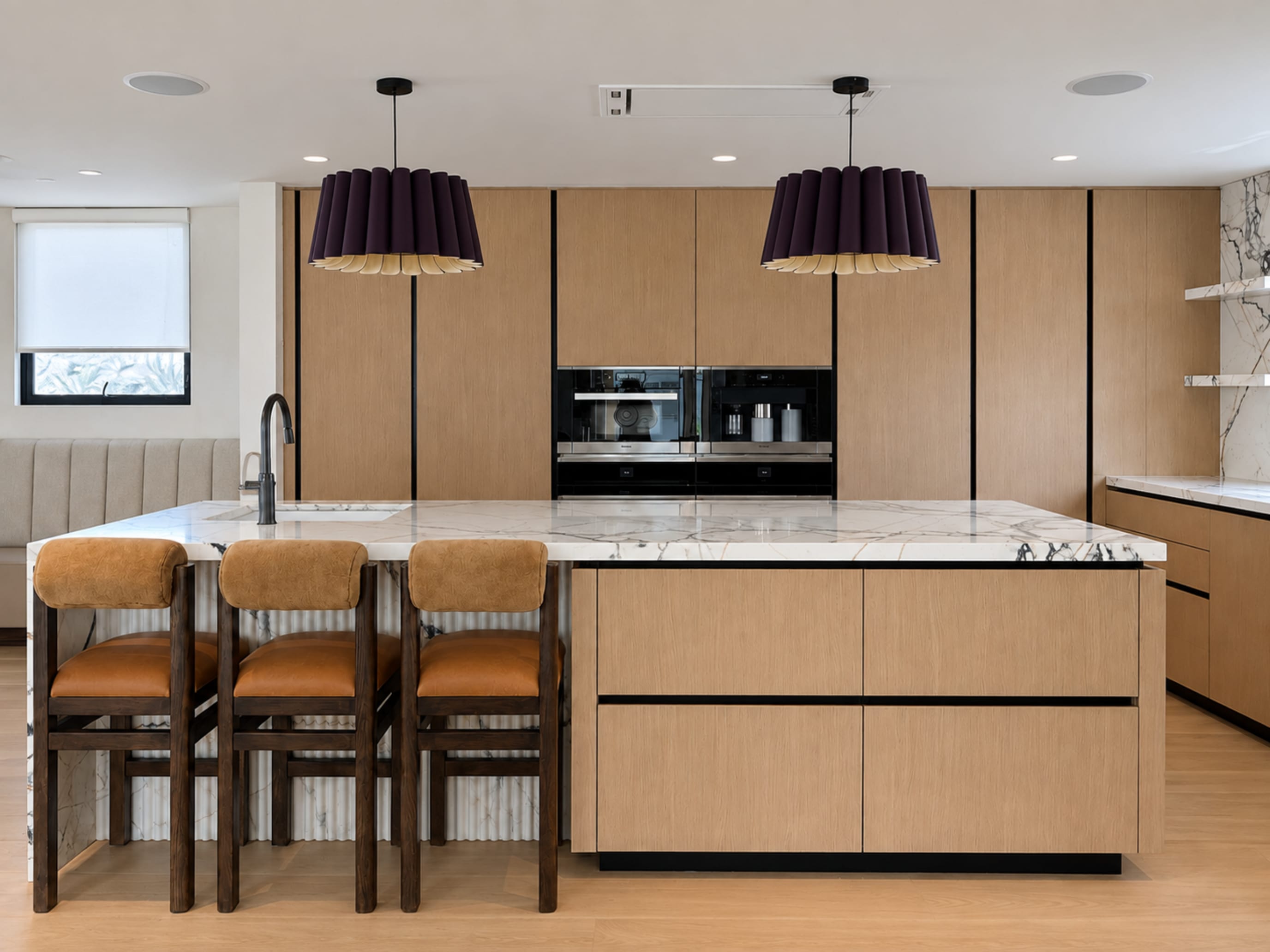 Oak kitchen cabinetry, veined stone island, leather counter stools, and dark sculptural pendant lights