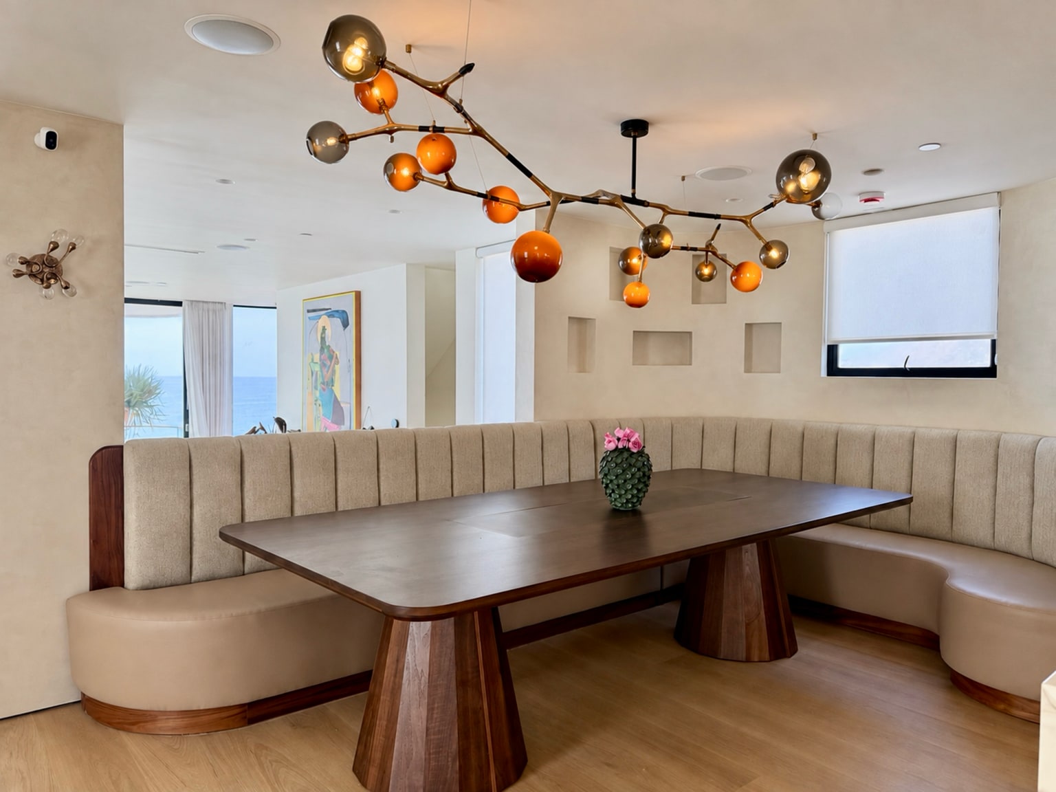 Walnut dining table beneath sculptural glass lighting beside a curved upholstered banquette, with the Pacific visible beyond