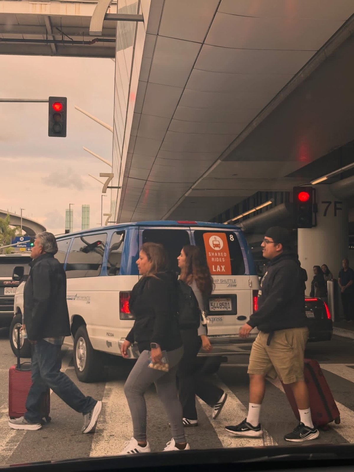 Passengers walking with luggage past a shuttle and traffic outside LAX Terminal 7