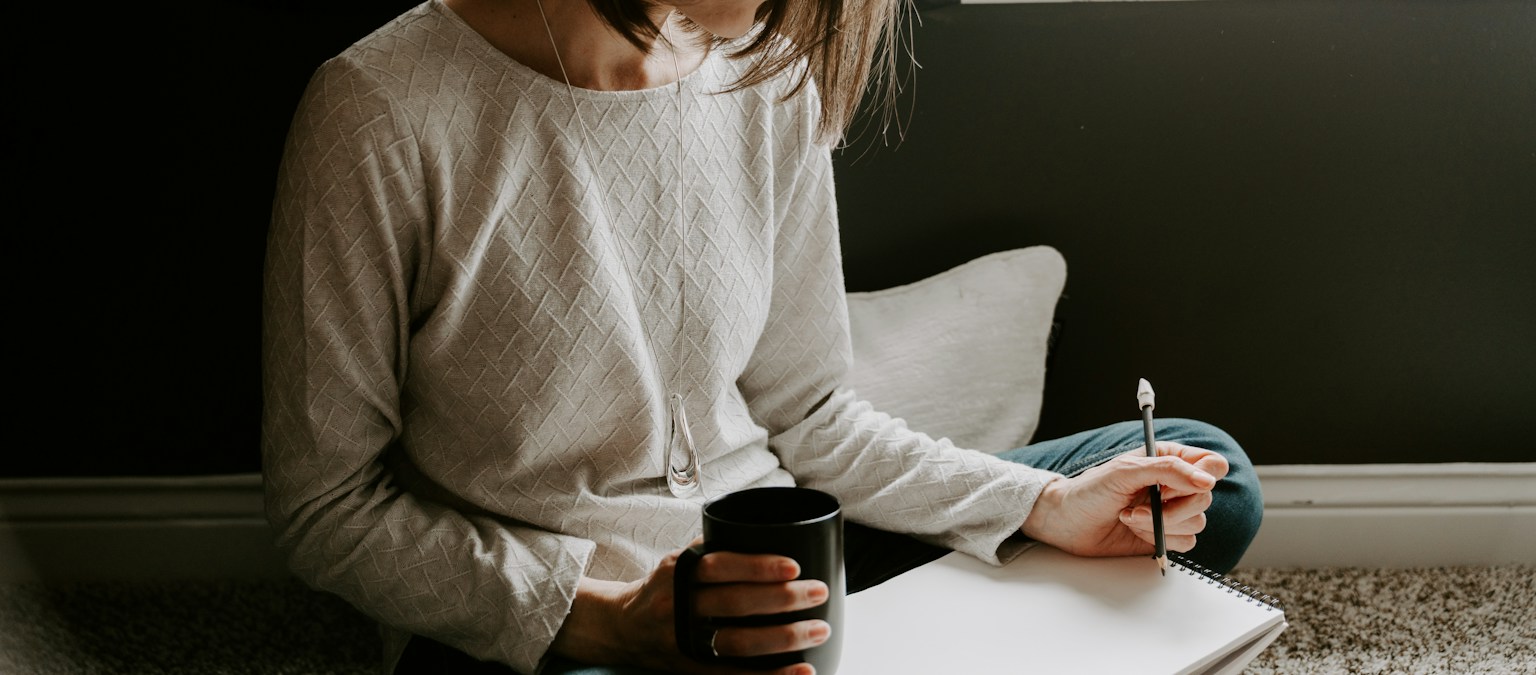 A woman writing in a notebook while holding a warm mug