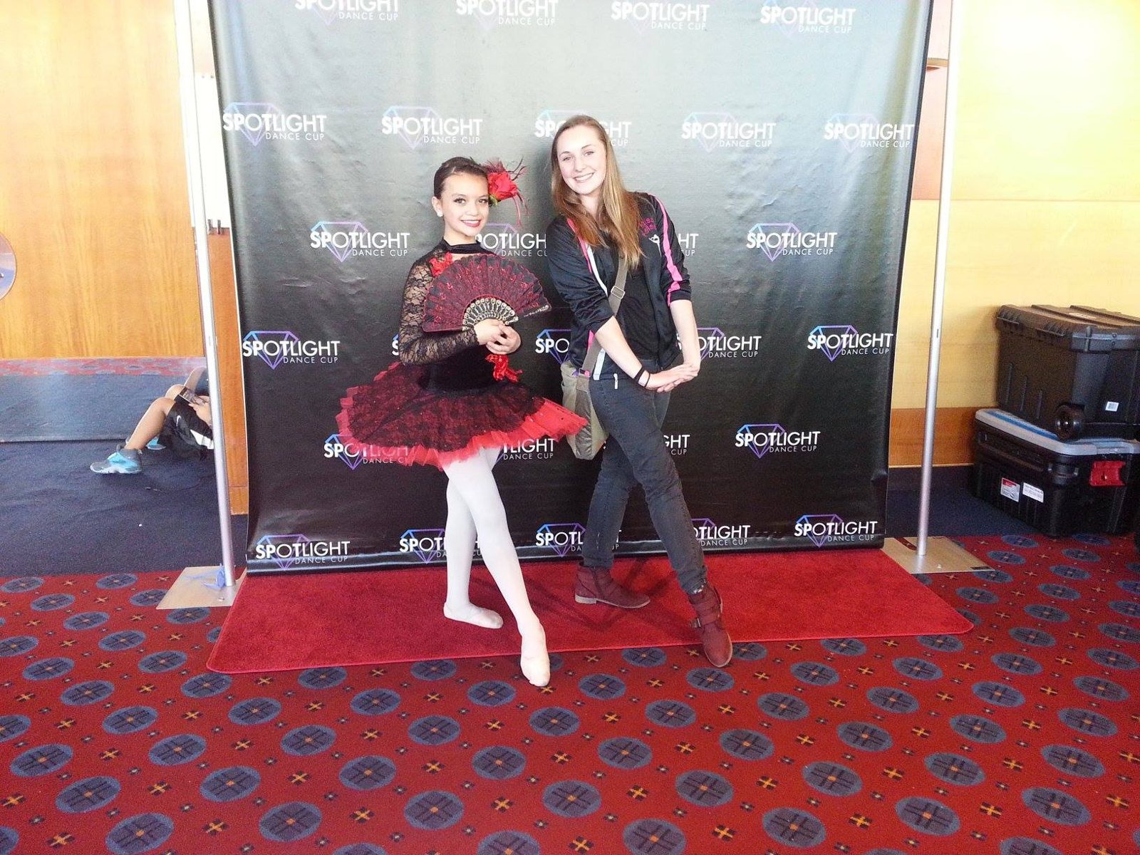 A young dancer in a red and black costume posing with an adult at a dance event