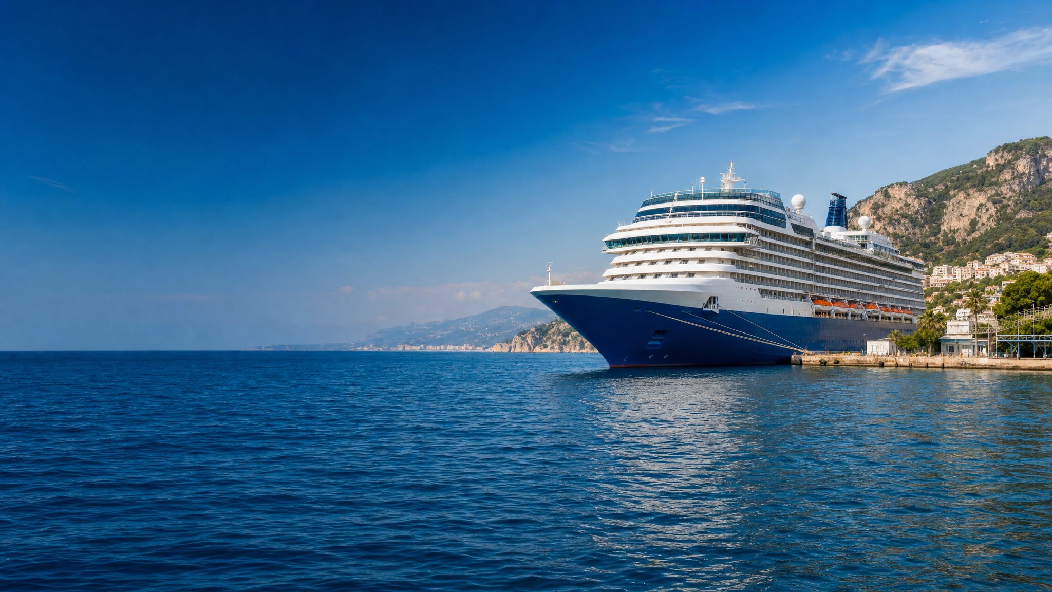A modern cruise ship docked in a bright Mediterranean harbor beneath coastal hills.