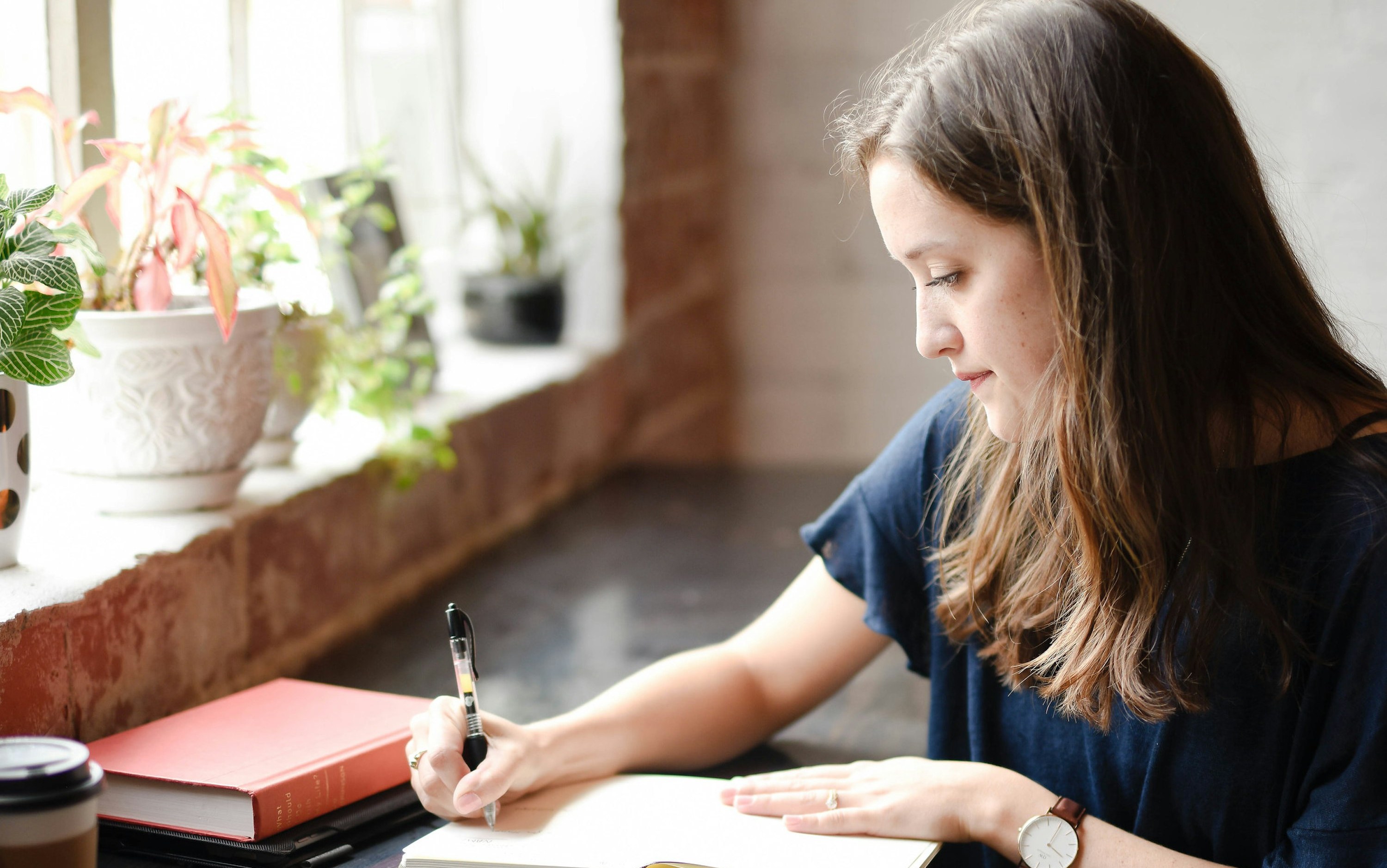 A woman writing thoughtfully in a notebook beside a window.