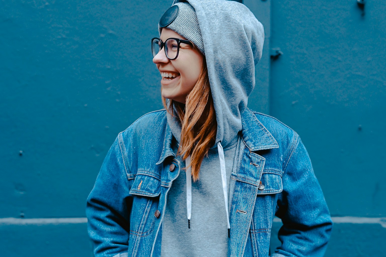 A woman in a gray hooded sweatshirt and denim jacket smiling against a blue wall.
