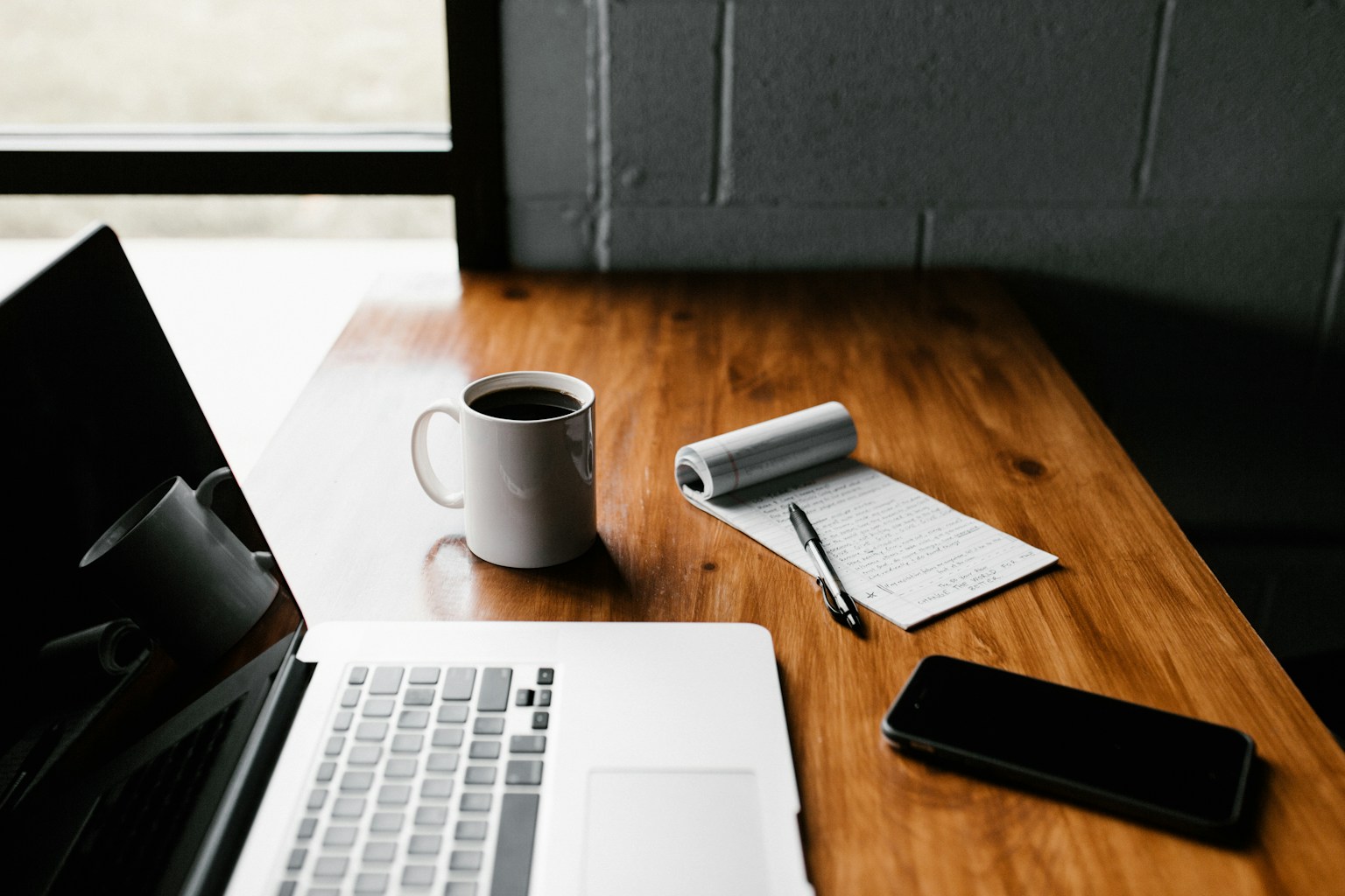 A phone, journal, pen, and mug on a wooden table near a window.