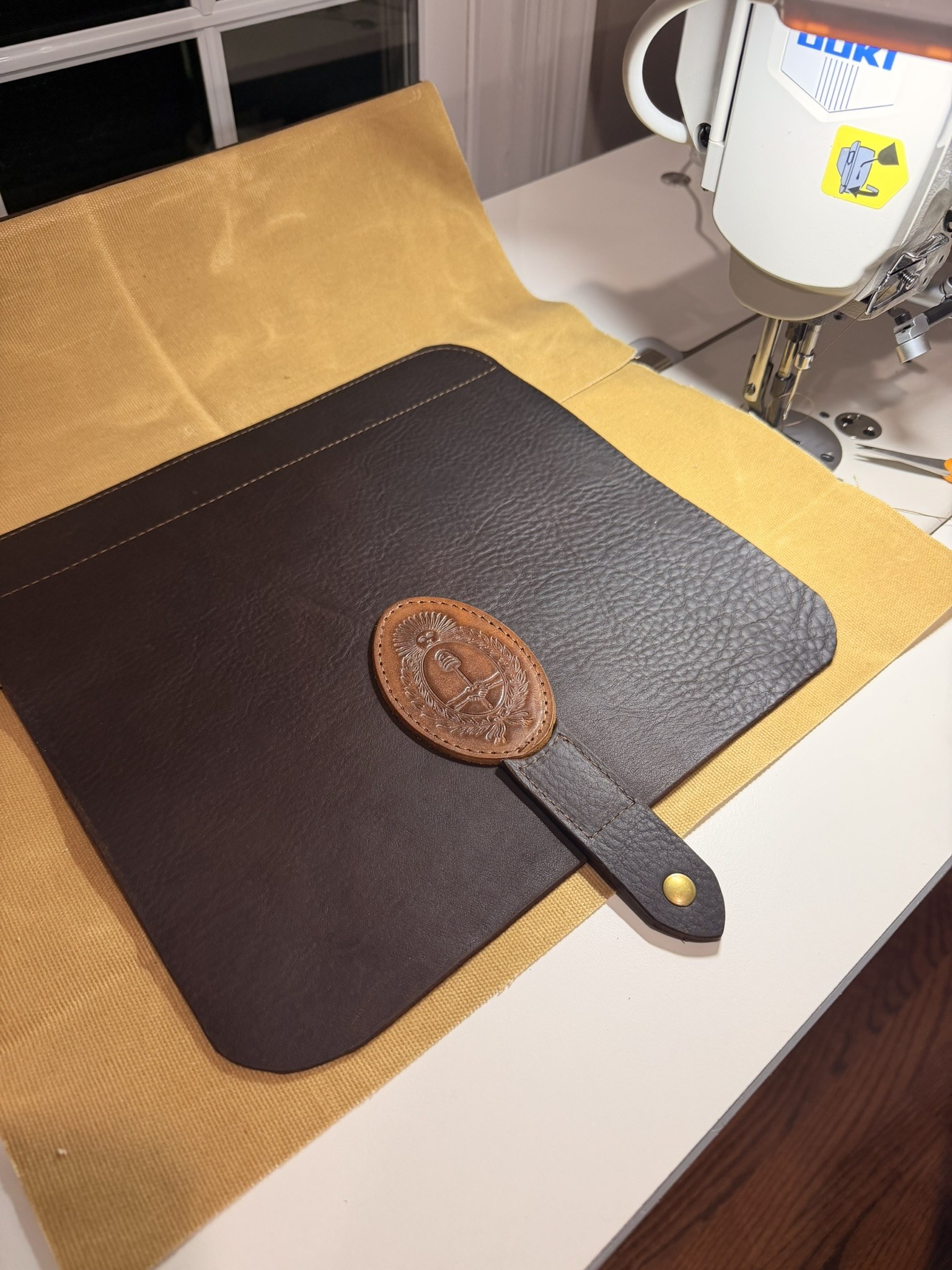 Brown leather shoulder bag panel with an Argentine coat of arms emblem on golden waxed canvas beside the workshop sewing machine