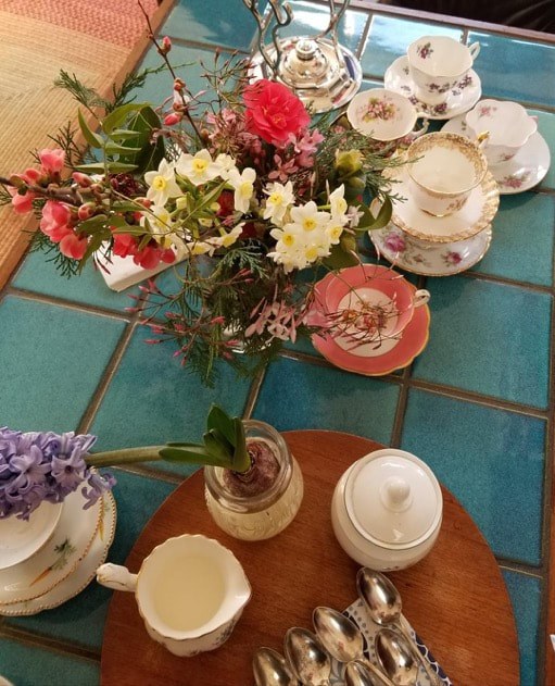 A colorful bouquet in a vase on a teal-tiled table with teacups and saucers.
