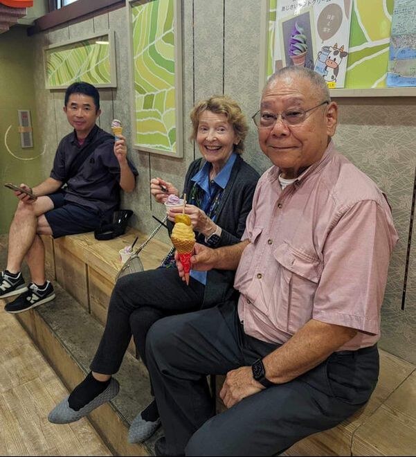 Three people sitting on a wooden bench, smiling and holding drinks.