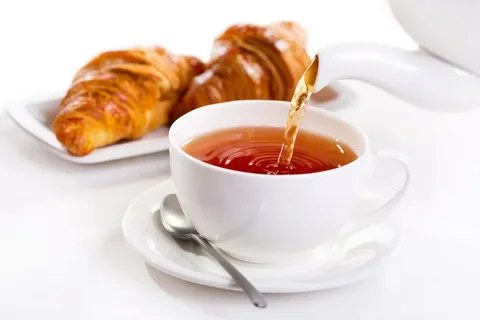 Tea being poured into a white cup with croissants in the background.