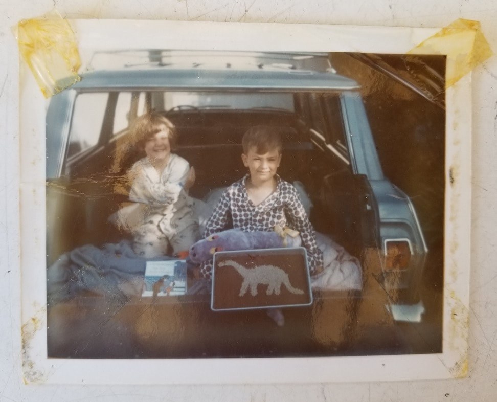 Two children sitting in the back of a vehicle with a dinosaur sign.