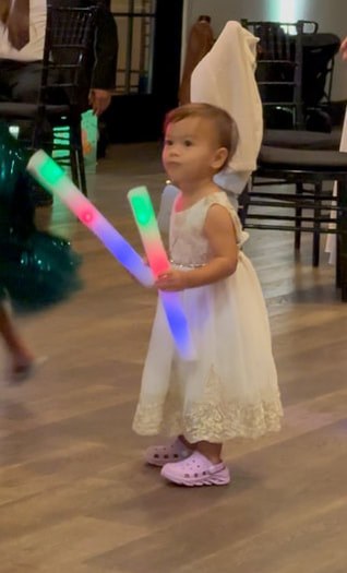 A toddler in a white dress holding colorful light sticks.