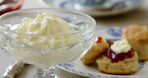 A glass dish filled with whipped cream beside a plate with scones and jam.