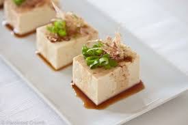 Three cubes of tofu garnished with green onions and bonito flakes on a white plate.