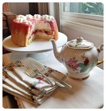 A layered cake with pink frosting and cream filling beside a floral teapot.
