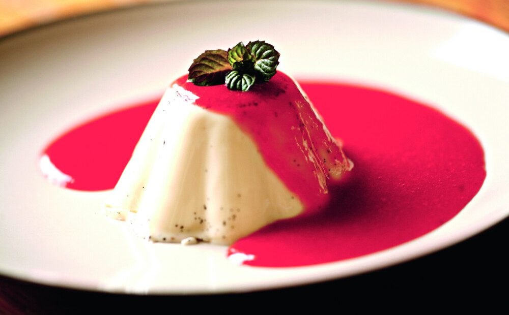 Creamy panna cotta dessert topped with a mint leaf, served with a vibrant berry sauce.