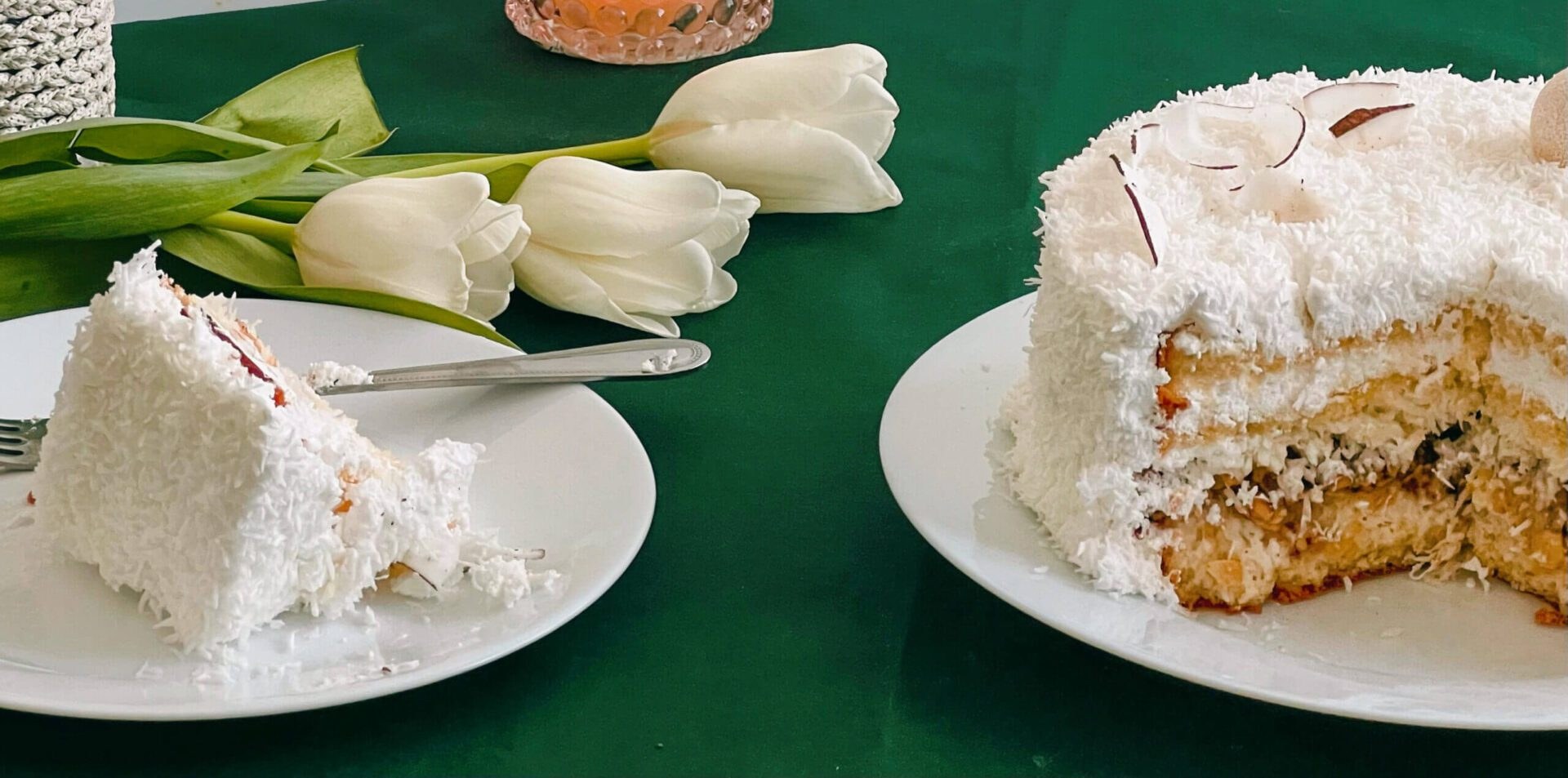 A slice of coconut cake on a plate beside the remaining cake on a green table.