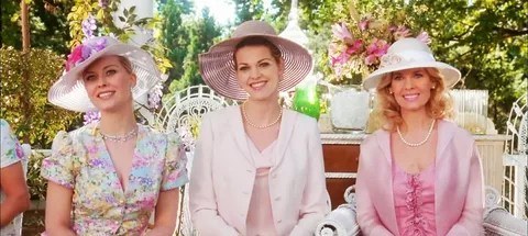A woman wearing a pink hat and light pink outfit smiles at an outdoor event.