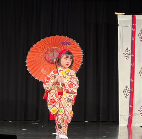 A young child in a traditional kimono holding a red parasol on stage.