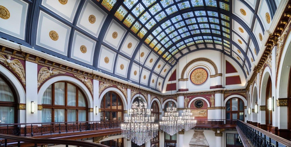 Elegant hall with arched windows and grand chandeliers under a glass ceiling.