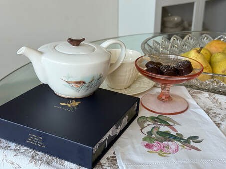 Elegant tea setup with a white teapot, glass dessert bowl, and floral tablecloth.