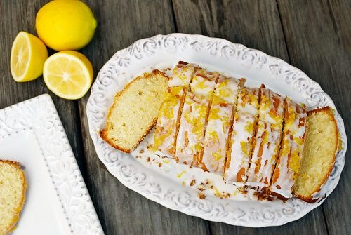 Lemon drizzle cake sliced on a white plate with lemons beside it.