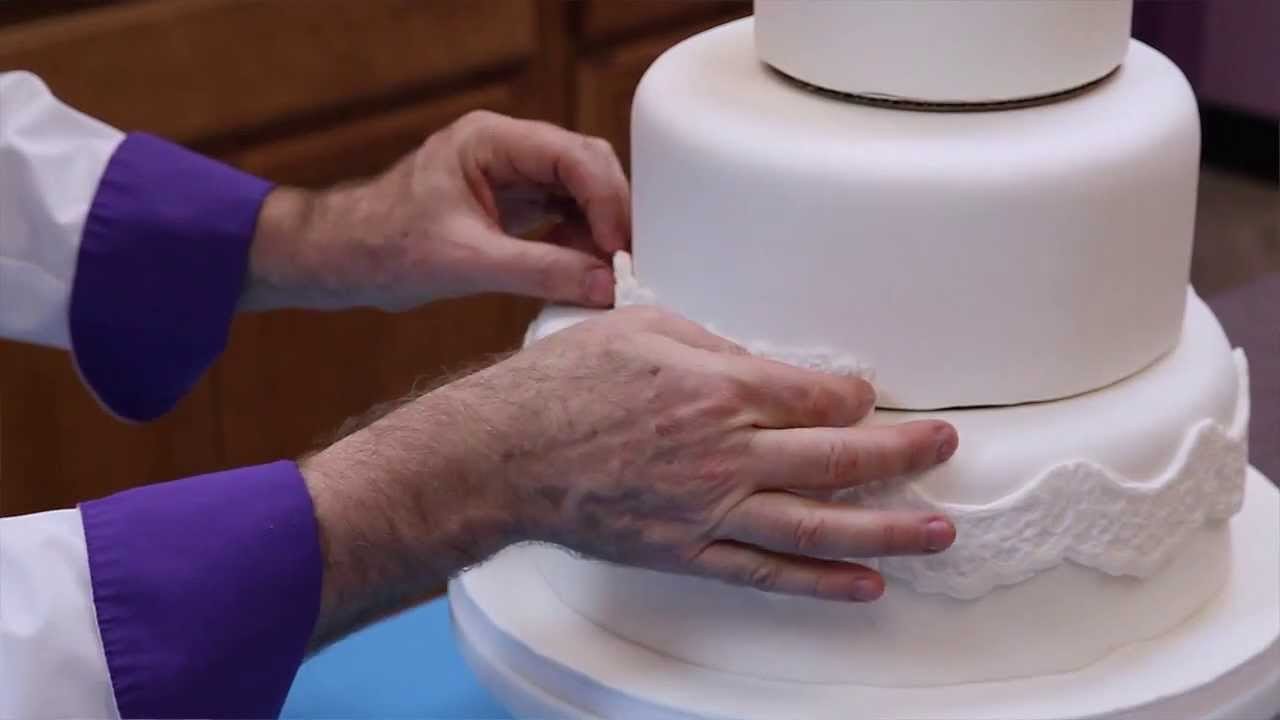 Hands decorating a white tiered cake with fondant or icing.