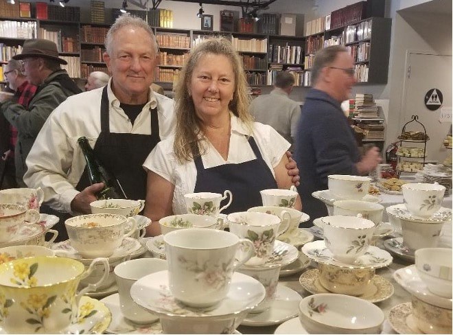 Two people smiling behind a table of fine china in a shop.
