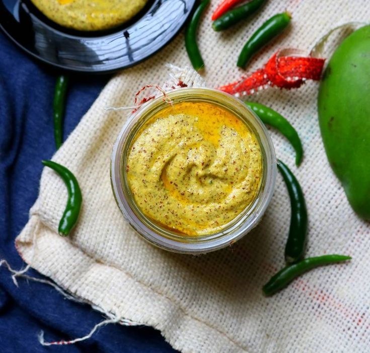 A jar of spicy green mango chutney surrounded by fresh chilies and a whole mango.