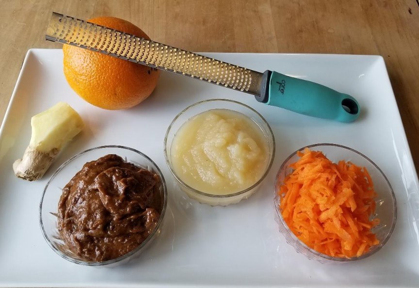 Three bowls with grated carrot, apple sauce, and chocolate spread, alongside a grater and an orange.