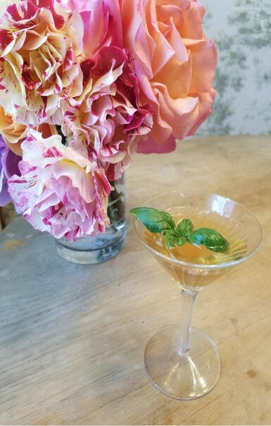 Elegant cocktail garnished with fresh basil beside a bouquet of pink flowers.