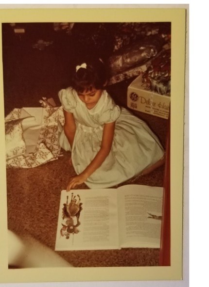 Young girl in a blue dress kneeling on the floor reading a book.