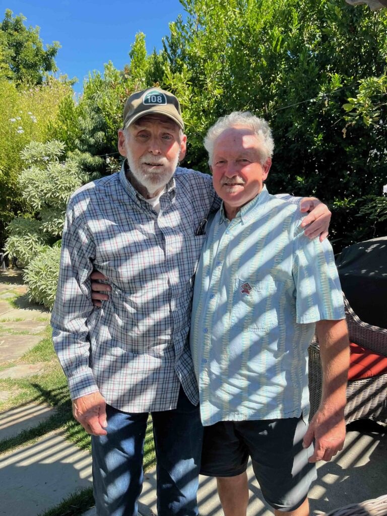 Two elderly men smiling outdoors with dappled sunlight.