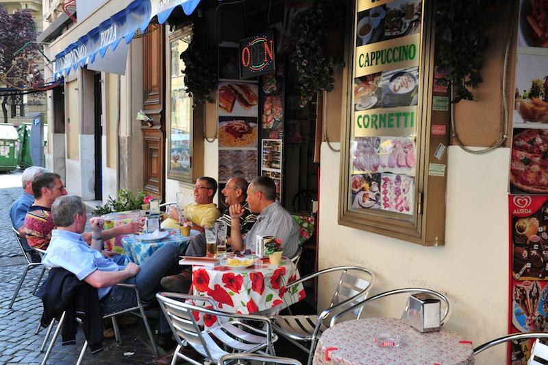 Two men enjoying drinks at an outdoor café with colorful tablecloths.