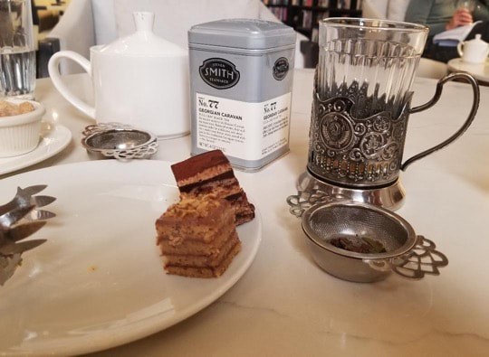 A layered chocolate cake slice on a plate next to an ornate glass and tea accessories.