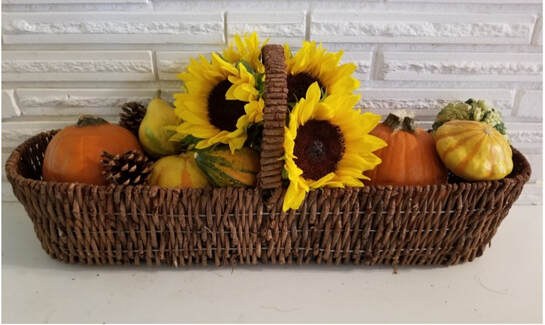 Sunflowers and small pumpkins in a woven basket against a white brick background.