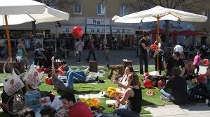 People relaxing on grass in a busy urban area with umbrellas and buildings.