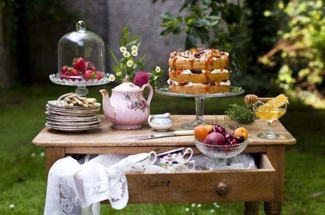 Rustic outdoor table set with cake, fruits, and tea items.