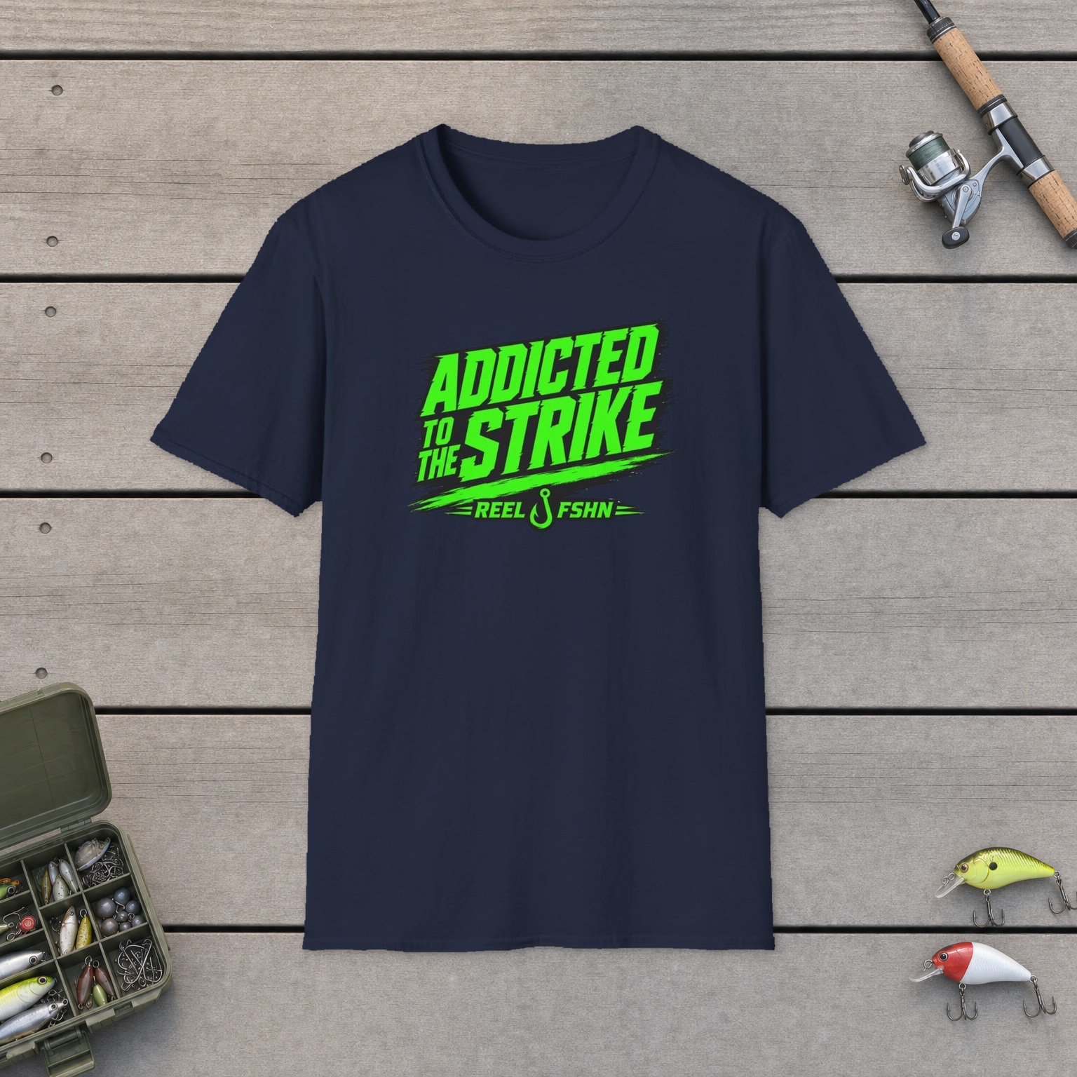Addicted To The Strike T-Shirt on a wooden dock with a fishing rod, reel, tackle box, and two lures