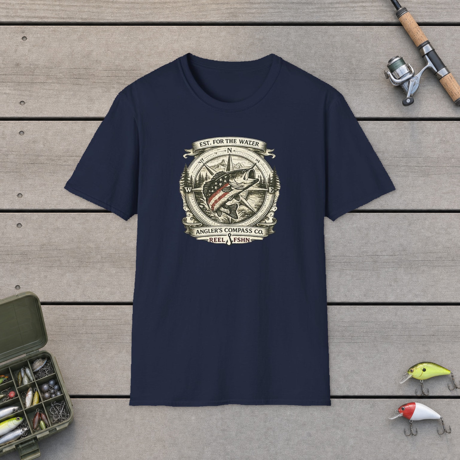 Angler's Compass Company T-Shirt on a wooden dock with a fishing rod, reel, tackle box, and two lures