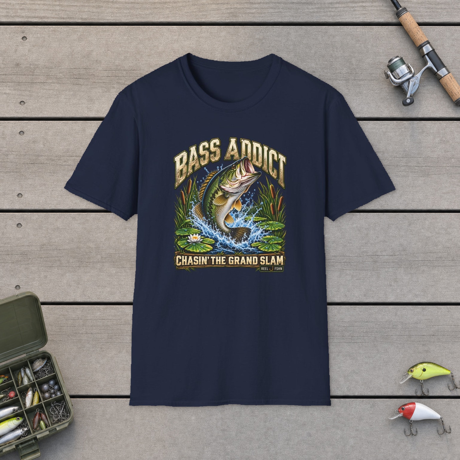 Bass Addict T-Shirt on a wooden dock with a fishing rod, reel, tackle box, and two lures