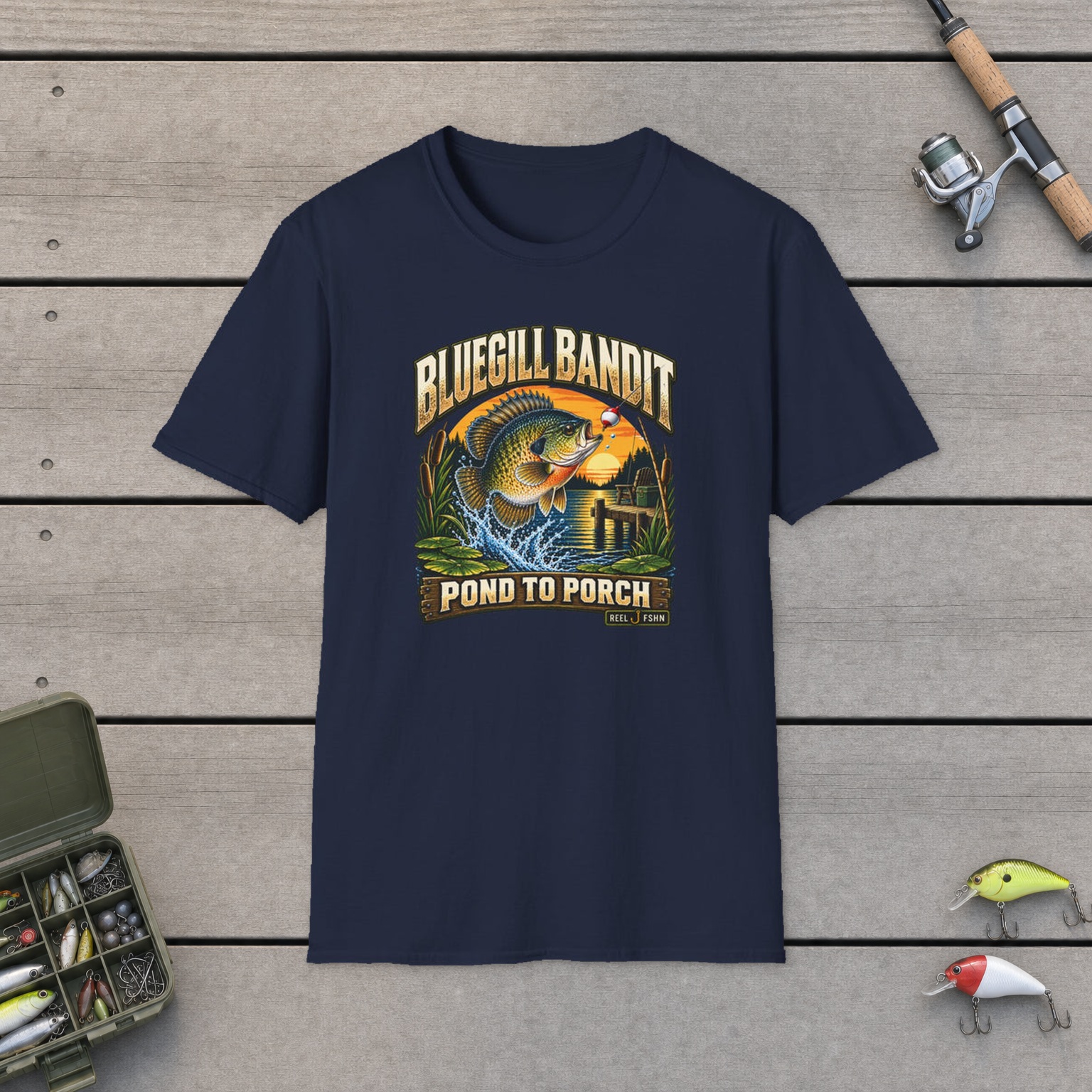 Bluegill Bandit T-Shirt on a wooden dock with a fishing rod, reel, tackle box, and two lures