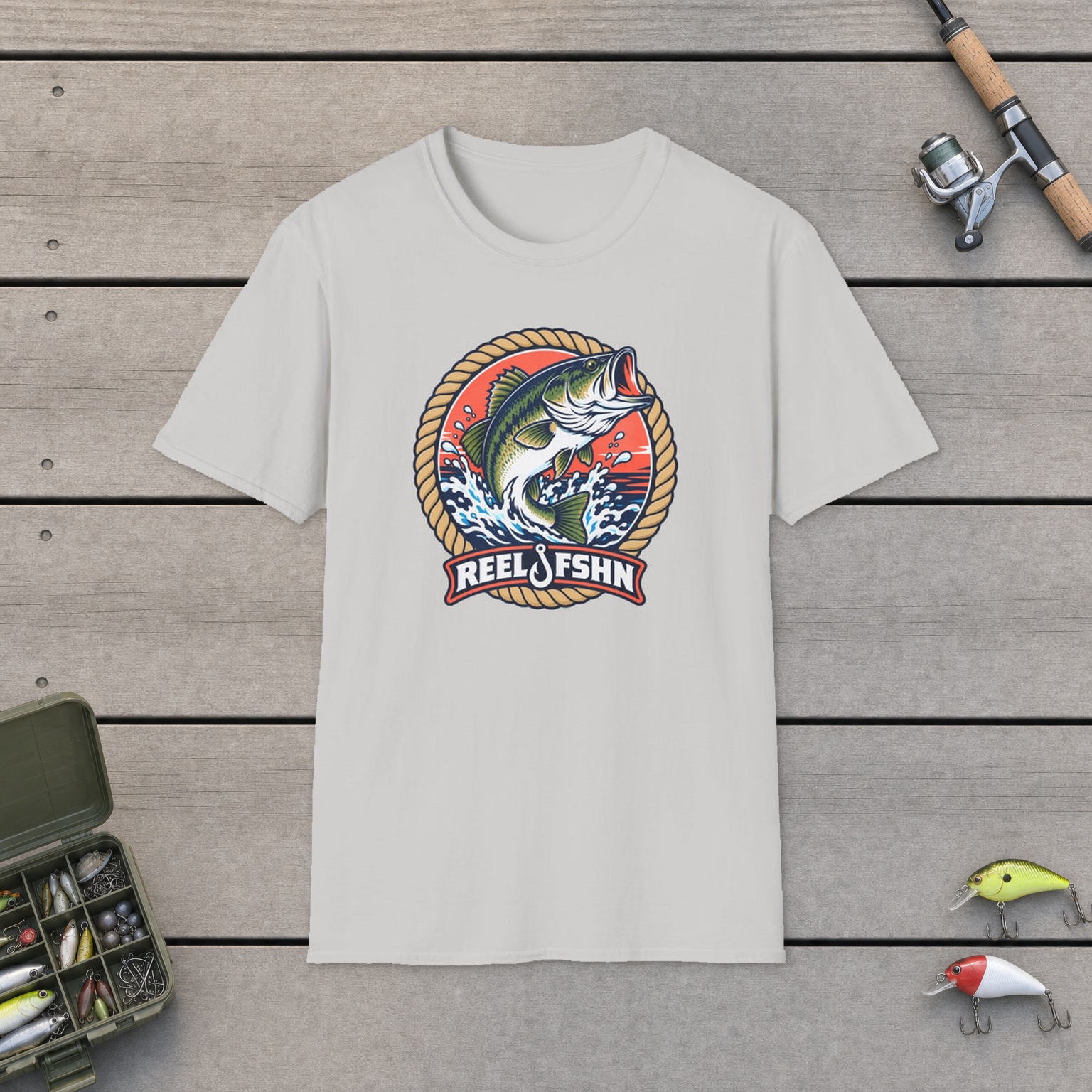 Coral Leaping Bass Badge T-Shirt on a wooden dock with a fishing rod, reel, tackle box, and two lures