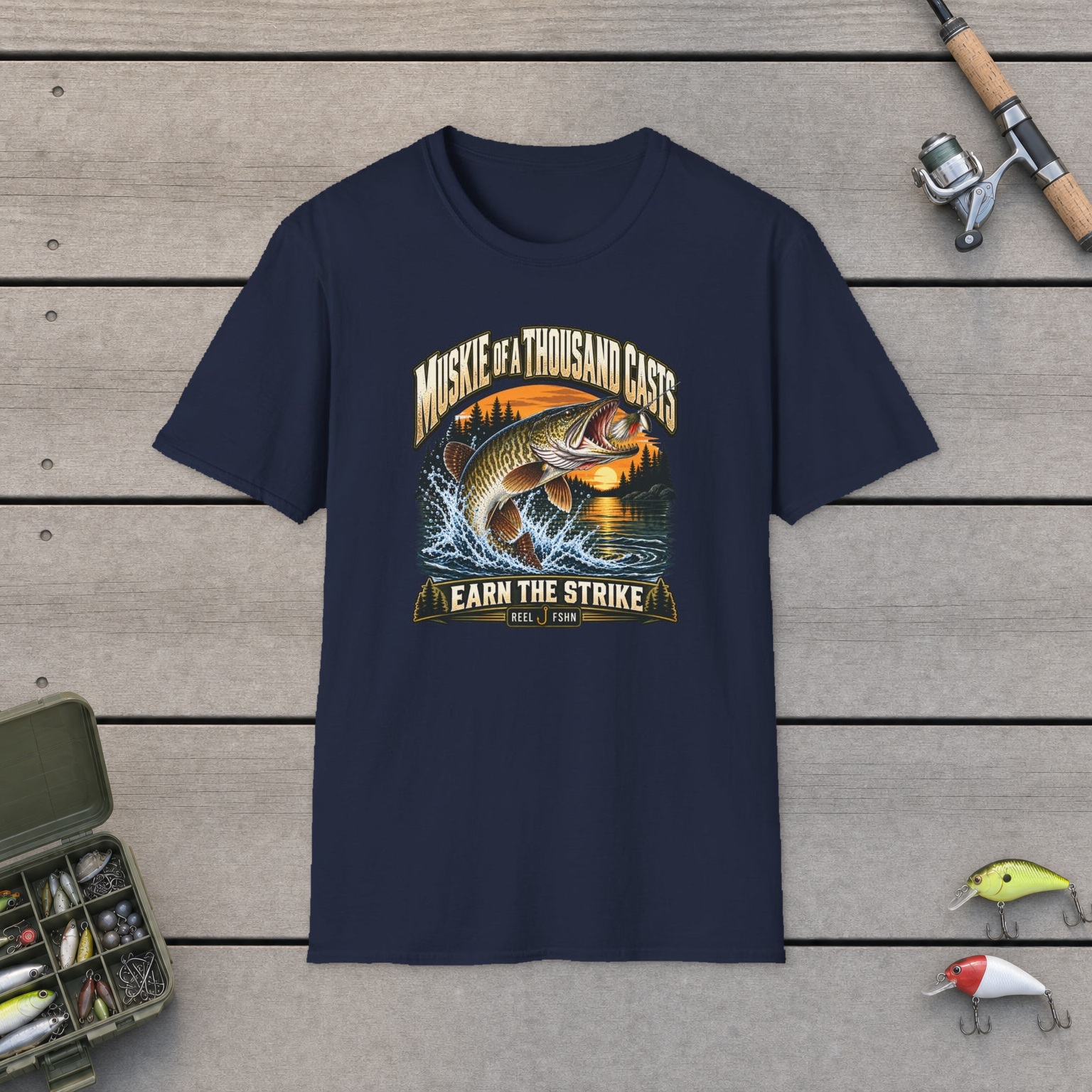 Muskie of a Thousand Casts T-Shirt on a wooden dock with a fishing rod, reel, tackle box, and two lures