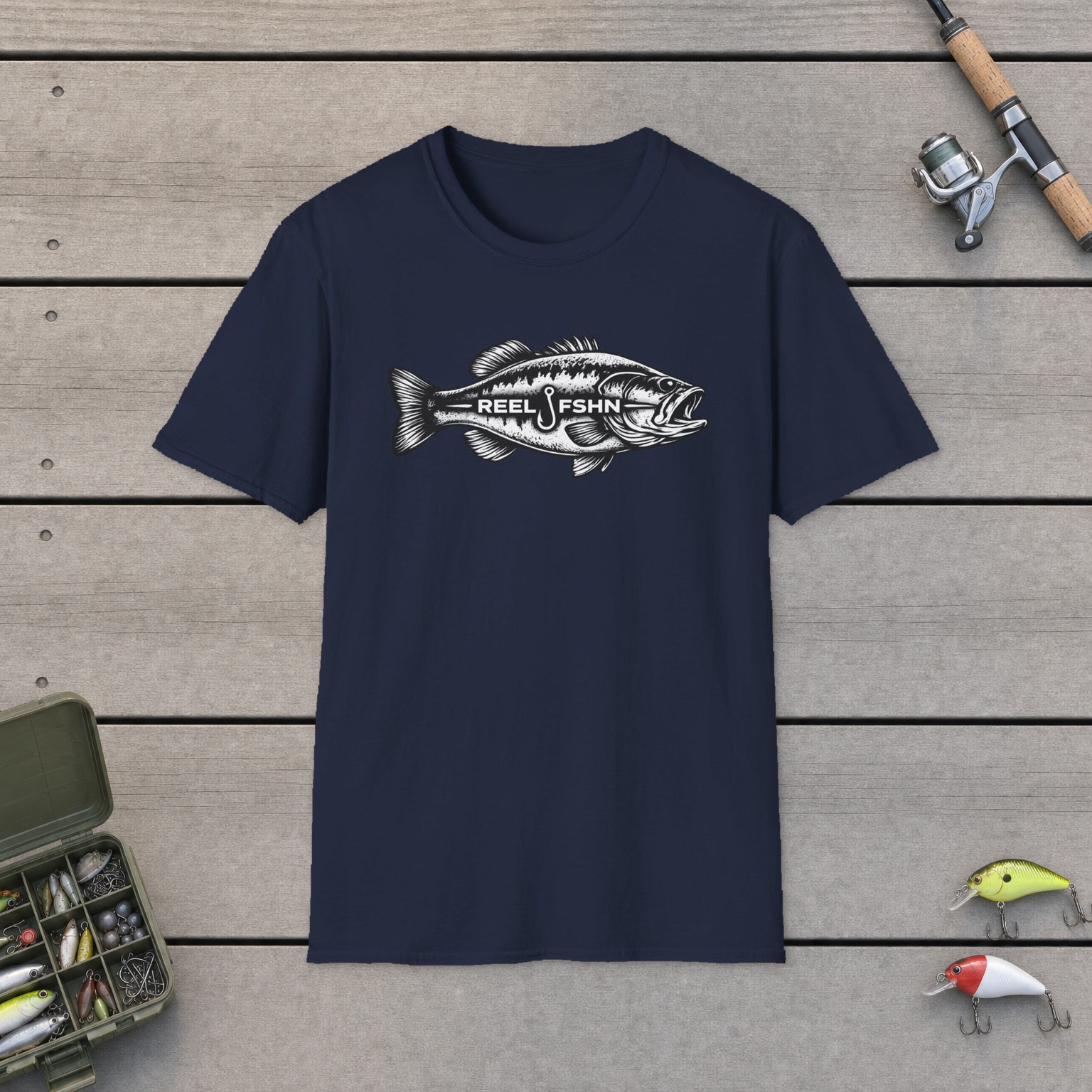 REEL FSHN BASS T-Shirt on a wooden dock with a fishing rod, reel, tackle box, and two lures