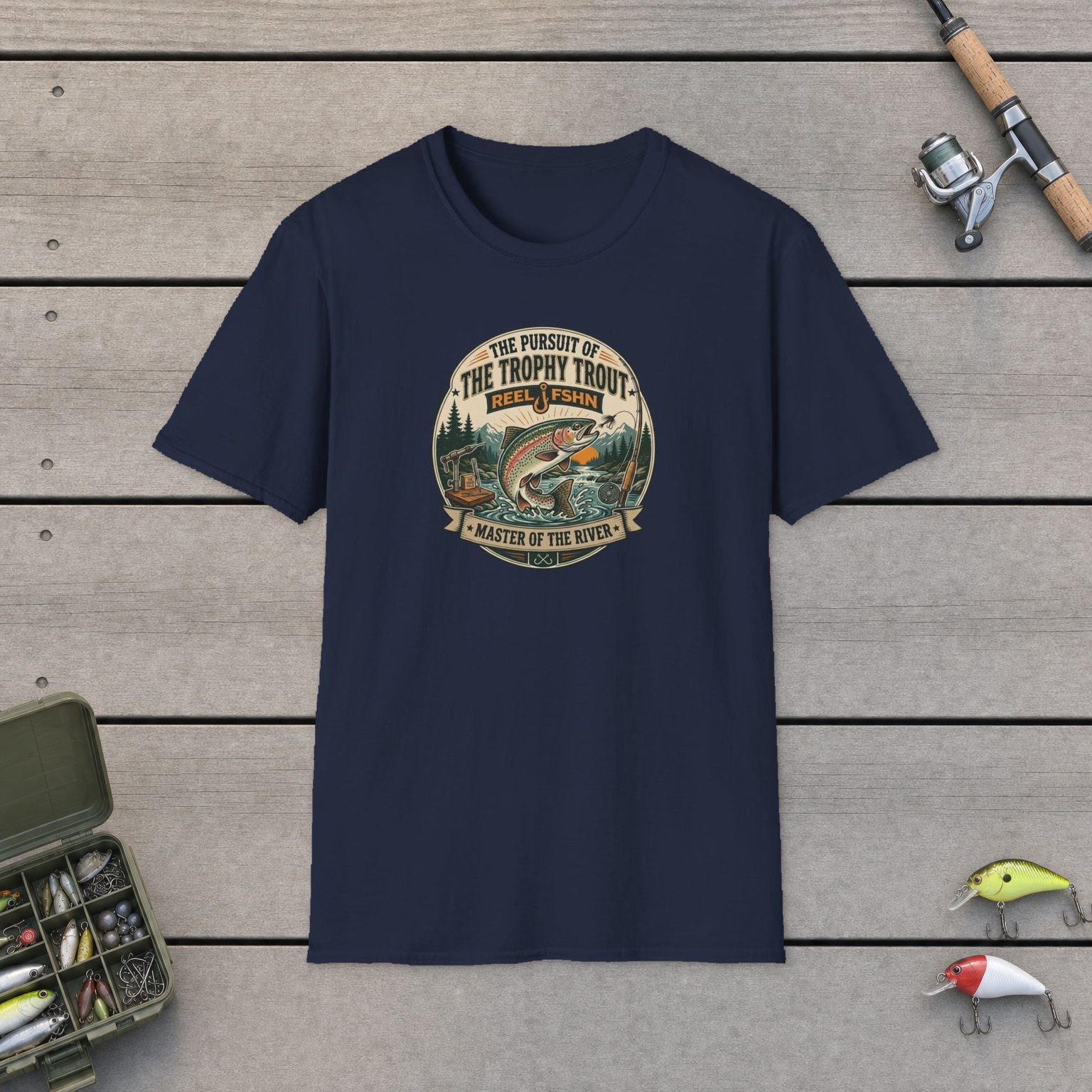 The Pursuit of The Trophy Trout T-Shirt on a wooden dock with a fishing rod, reel, tackle box, and two lures