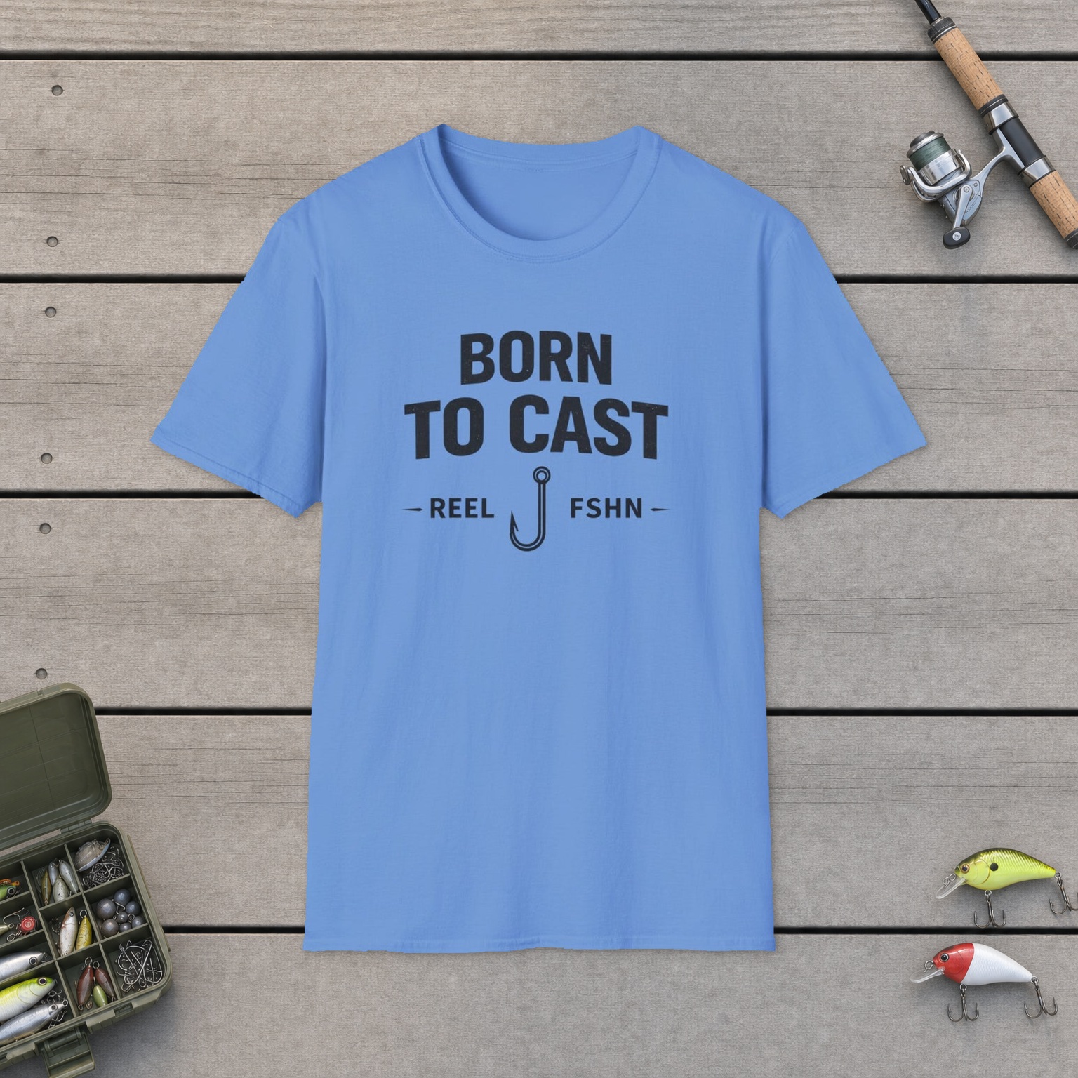 Born to Cast Fishing T-Shirt on a wooden dock with a fishing rod, reel, tackle box, and two lures