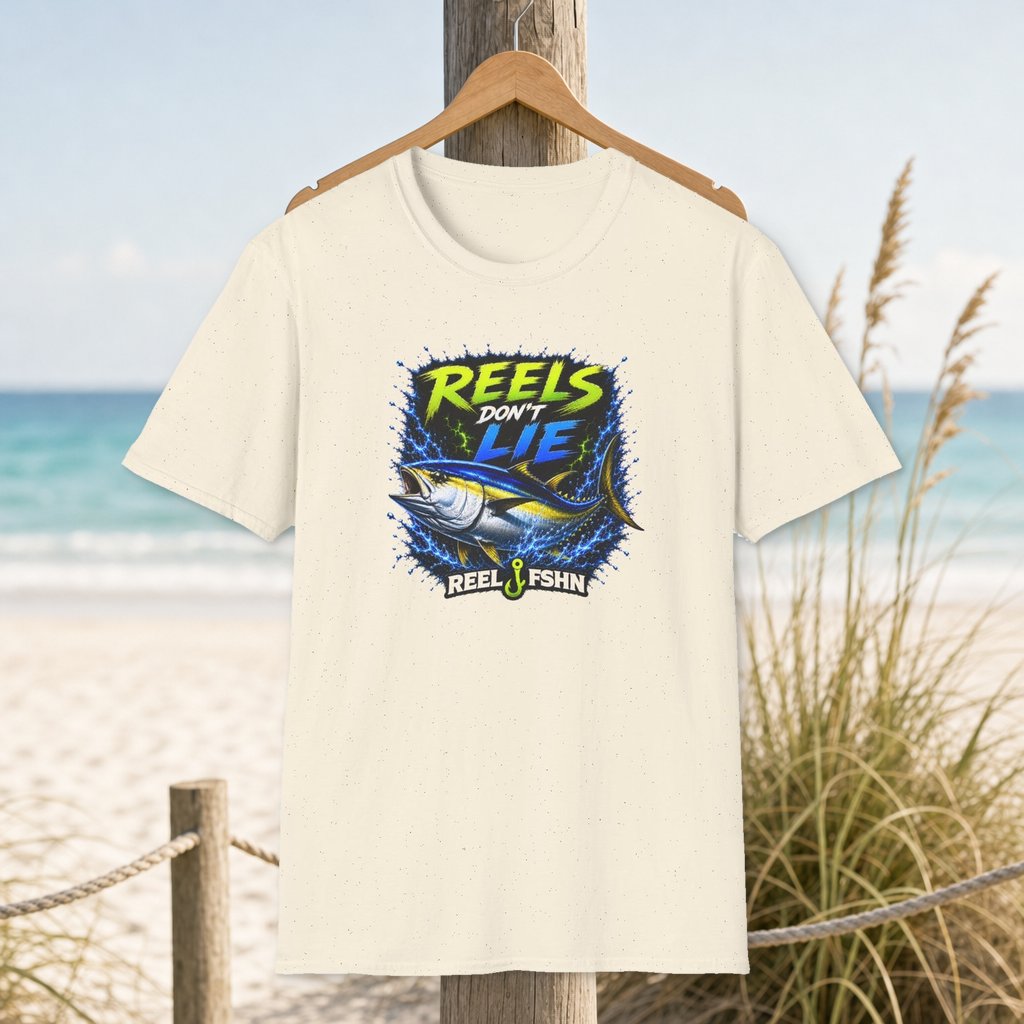 Reels Don't Lie T-Shirt on a hanger beside a sandy beach and turquoise coastal water